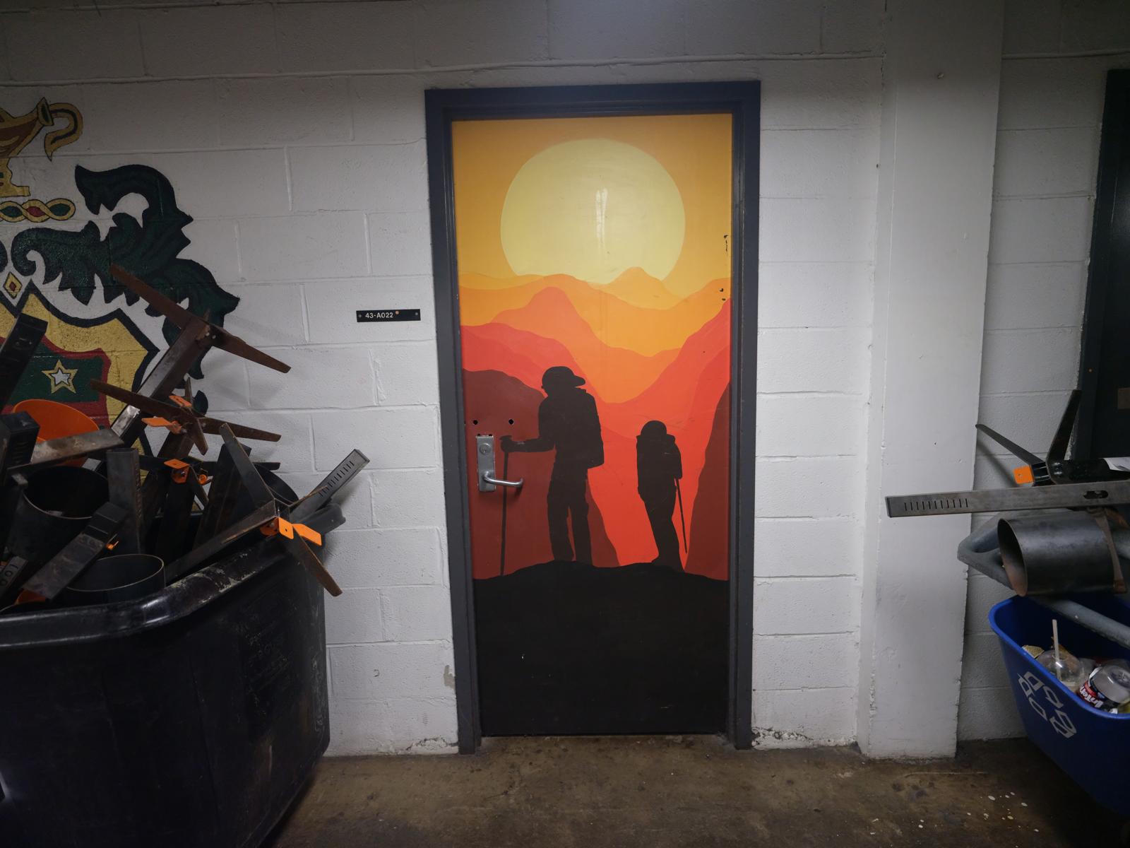A door with a painting of two silhouetted people hiking on a hill, with rolling
hills and a sunset in the background.